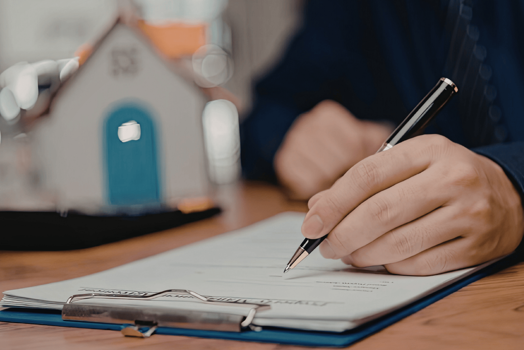 A close-up of a hand signing papers on a clipboard with a small house model in the background