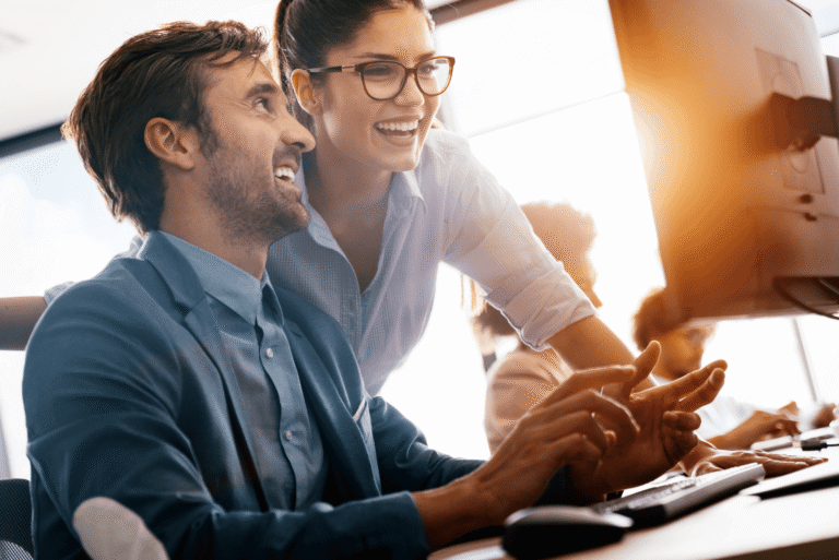 Two coworkers smiling and looking at a computer screen together in a bright office.
